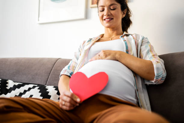 femme enceinte souriante, assise en teilleur sur un canapé gris et tenant un coeur rouge en papaier dans la main doite l'autre main se trouvant au dessus de son ventre bien arrondi